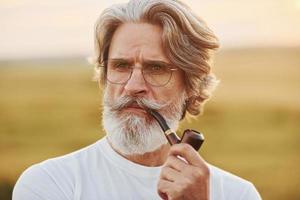 retrato de un anciano elegante con cabello gris y barba que está parado al aire libre en el campo en un día soleado y fumando foto