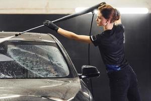 Using equipment with high pressure water. Modern black automobile get cleaned by woman inside of car wash station