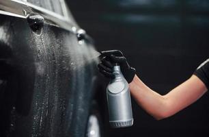 rociando el vehículo. el automóvil negro moderno es limpiado por una mujer dentro de la estación de lavado de autos foto