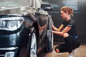 Wipes surface by using towel. Modern black automobile get cleaned by woman inside of car wash station photo