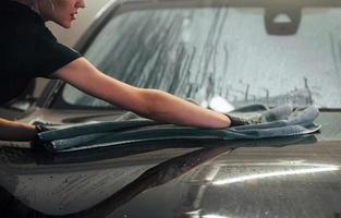 Wipes surface by using towel. Modern black automobile get cleaned by woman inside of car wash station photo