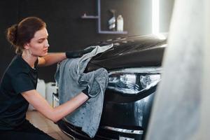 Wipes surface by using towel. Modern black automobile get cleaned by woman inside of car wash station photo