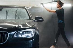 Using equipment with high pressure water. Modern black automobile get cleaned by woman inside of car wash station