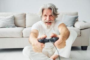 juega videojuegos usando el controlador. Senior hombre moderno con estilo con pelo gris y barba en el interior foto