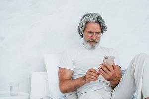 Sits on bed and using phone. Senior stylish modern man with grey hair and beard indoors photo