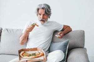 Eats delicious pizza while watching TV show. Senior stylish modern man with grey hair and beard indoors photo