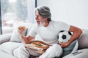 Soccer fan watching match and eats pizza. Senior stylish modern man with grey hair and beard indoors photo