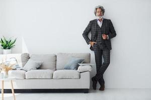 en ropa elegante. de pie contra la pared blanca. Senior hombre moderno con estilo con pelo gris y barba en el interior foto