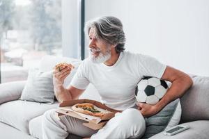 Soccer fan watching match and eats pizza. Senior stylish modern man with grey hair and beard indoors photo