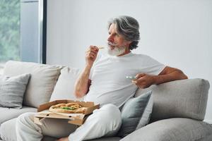 Eats delicious pizza while watching TV show. Senior stylish modern man with grey hair and beard indoors photo