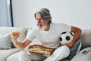 Soccer fan watching match and eats pizza. Senior stylish modern man with grey hair and beard indoors photo