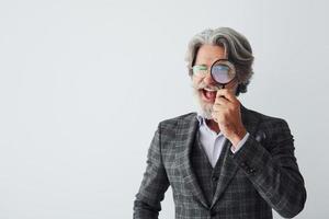 Having fun with magnifying glass. Senior stylish modern man with grey hair and beard indoors photo