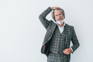 In elegant clothes. Standing against white wall. Senior stylish modern man with grey hair and beard indoors photo