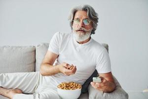 Watching TV show and eats popcorn. Senior stylish modern man with grey hair and beard indoors photo