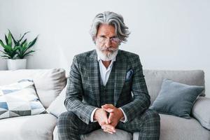 Sits on comfortable sofa in formal clothes. Senior stylish modern man with grey hair and beard indoors photo