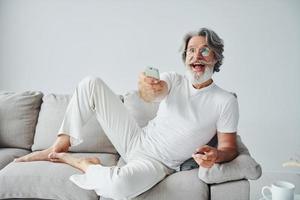 se sienta en el sofá. Senior hombre moderno con estilo con pelo gris y barba en el interior foto