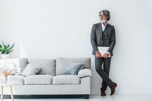 hombre moderno con estilo senior con pelo gris y barba en el interior de pie cerca de la pared con el portátil en las manos foto