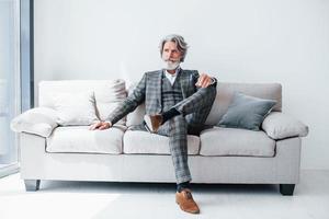 Sits on comfortable sofa in formal clothes. Senior stylish modern man with grey hair and beard indoors photo