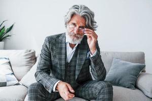 Sits on comfortable sofa in formal clothes. Senior stylish modern man with grey hair and beard indoors photo