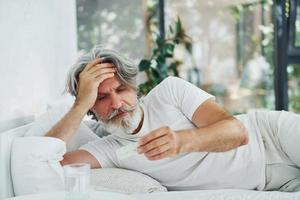 Sick senior stylish modern man with grey hair and beard indoors lying on the bed and feels bad photo