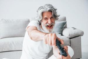 juega videojuegos usando el controlador. Senior hombre moderno con estilo con pelo gris y barba en el interior foto