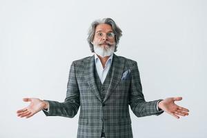 en ropa elegante. de pie contra la pared blanca. Senior hombre moderno con estilo con pelo gris y barba en el interior foto