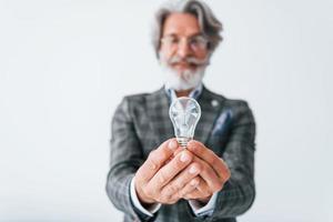 With light bulb in hands. Senior stylish modern man with grey hair and beard indoors photo