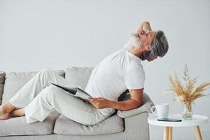 Sits on the sofa. Senior stylish modern man with grey hair and beard indoors photo