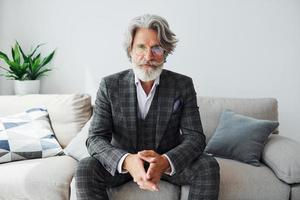 Sits on comfortable sofa in formal clothes. Senior stylish modern man with grey hair and beard indoors photo