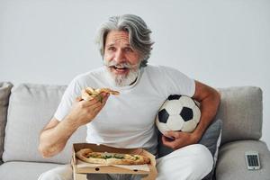 Soccer fan watching match and eats pizza. Senior stylish modern man with grey hair and beard indoors photo