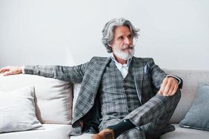 Sits on comfortable sofa in formal clothes. Senior stylish modern man with grey hair and beard indoors photo