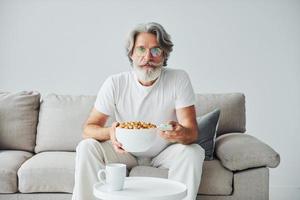 viendo un programa de televisión y come palomitas de maíz. Senior hombre moderno con estilo con pelo gris y barba en el interior foto