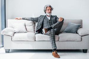 Sits on comfortable sofa in formal clothes. Senior stylish modern man with grey hair and beard indoors photo