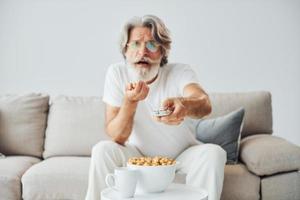 Watching TV show and eats popcorn. Senior stylish modern man with grey hair and beard indoors photo