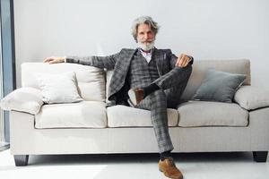 Sits on comfortable sofa in formal clothes. Senior stylish modern man with grey hair and beard indoors photo