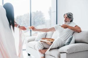 viendo la televisión con una mujer aterradora con el pelo negro cerca de él. Senior hombre moderno con estilo con pelo gris y barba en el interior foto