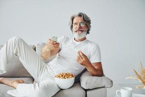 Watching TV show and eats popcorn. Senior stylish modern man with grey hair and beard indoors photo