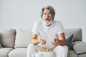 Watching TV show and eats popcorn. Senior stylish modern man with grey hair and beard indoors photo