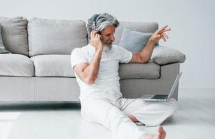 se sienta en el suelo cerca del sofá con una computadora portátil y escucha música. Senior hombre moderno con estilo con pelo gris y barba en el interior foto