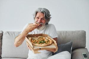 Eats delicious pizza while watching TV show. Senior stylish modern man with grey hair and beard indoors photo