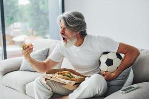 Soccer fan watching match and eats pizza. Senior stylish modern man with grey hair and beard indoors photo