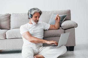 se sienta en el suelo cerca del sofá con una computadora portátil y escucha música. Senior hombre moderno con estilo con pelo gris y barba en el interior foto