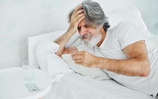 Sick senior stylish modern man with grey hair and beard indoors lying on the bed and feels bad photo