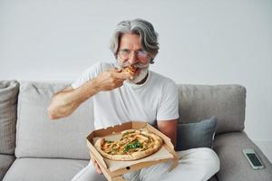 Eats delicious pizza while watching TV show. Senior stylish modern man with grey hair and beard indoors photo