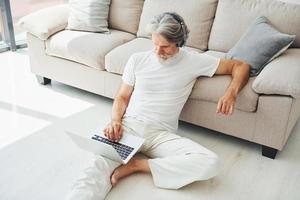 Sits on the ground near sofa with laptop and listens music. Senior stylish modern man with grey hair and beard indoors photo