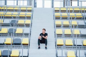 Sits with bottle of water. Sportive young guy in black shirt and pants outdoors at daytime photo