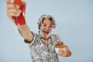 Bottle of drink and popcorn in hands. Senior stylish modern man with grey hair and beard indoors photo