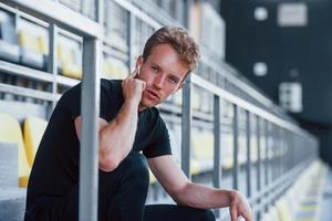 Listens to music when sitting. Sportive young guy in black shirt and pants outdoors at daytime photo