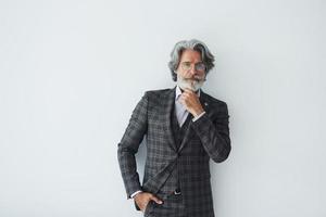 posando para una cámara mientras está de pie contra un fondo blanco. Senior hombre moderno con estilo con pelo gris y barba en el interior foto