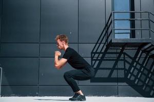 Exercising near building with black wall. Sportive young guy in black shirt and pants outdoors at daytime photo